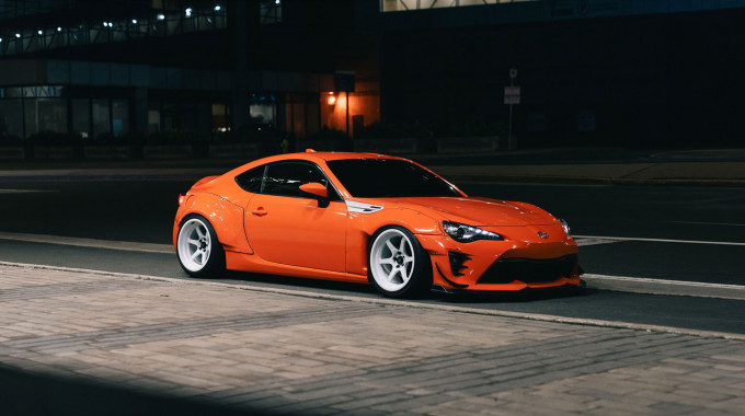Orange widebody Toyota sports coupe with white aftermarket wheels and a lowered stance, parked on a city street at night, an example of a modified car